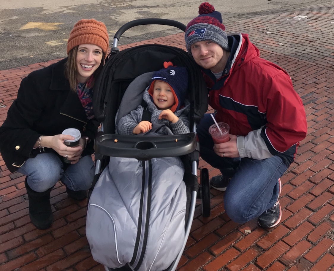 Sara and her husband squat and smile next to their son in a stroller