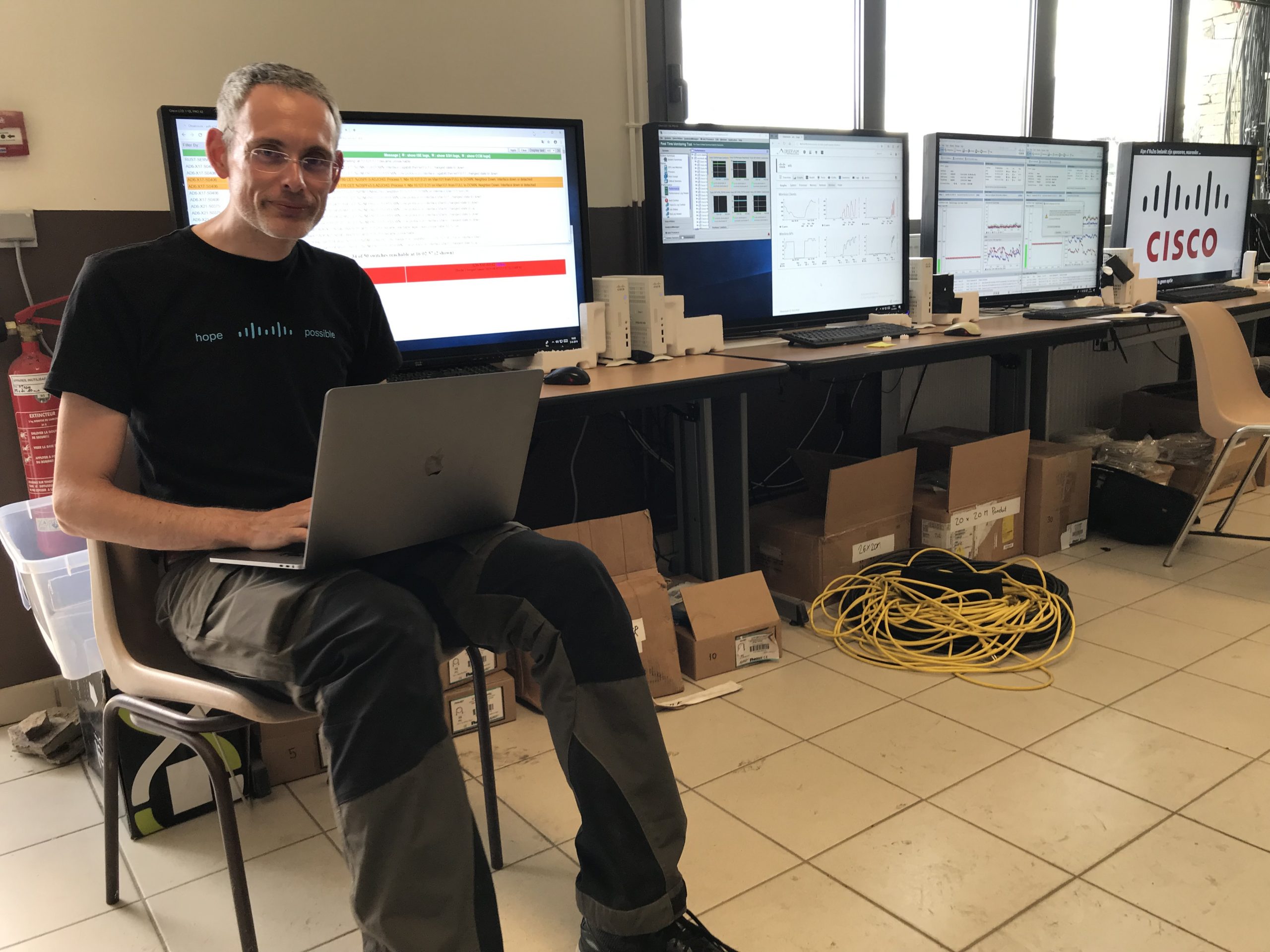 Mark sits with his laptop on his lap and a series of large monitors behind him at the network operating center for the Alpe d’HuZes event.
