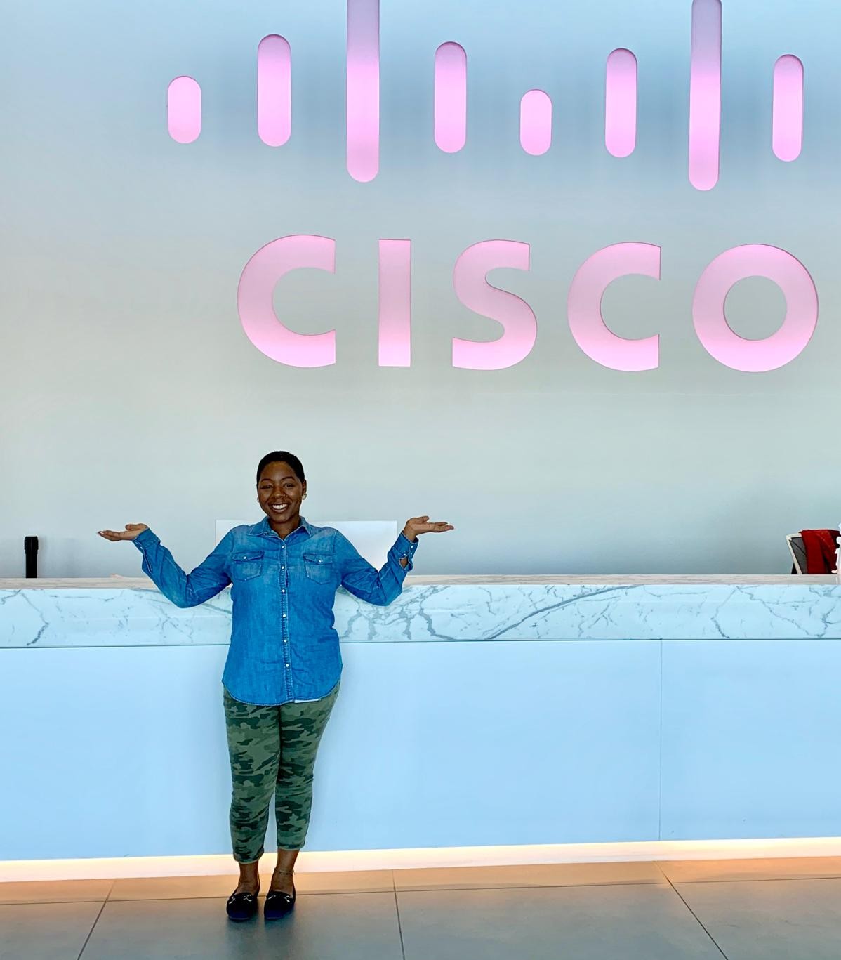 Amber stands in front of a bright pink light up Cisco sign in a lobby of a building.