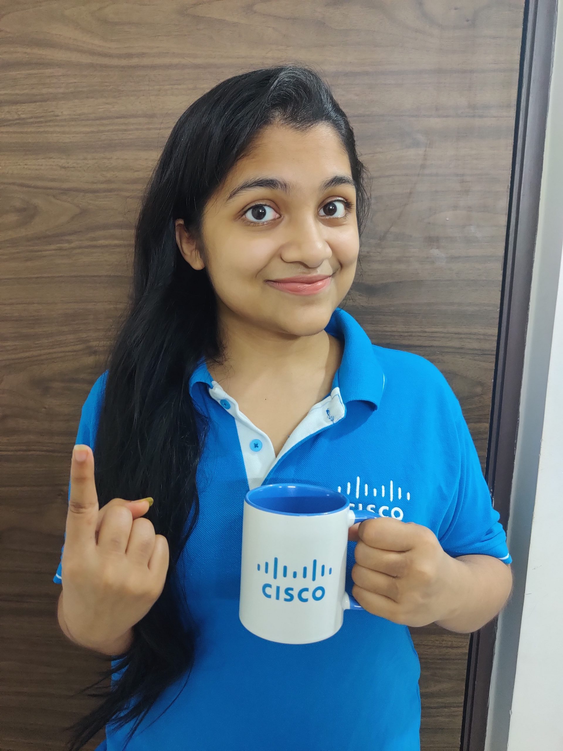 Diparati wears a Cisco shirt while holding a cup of coffee and holding up her finger as #1.