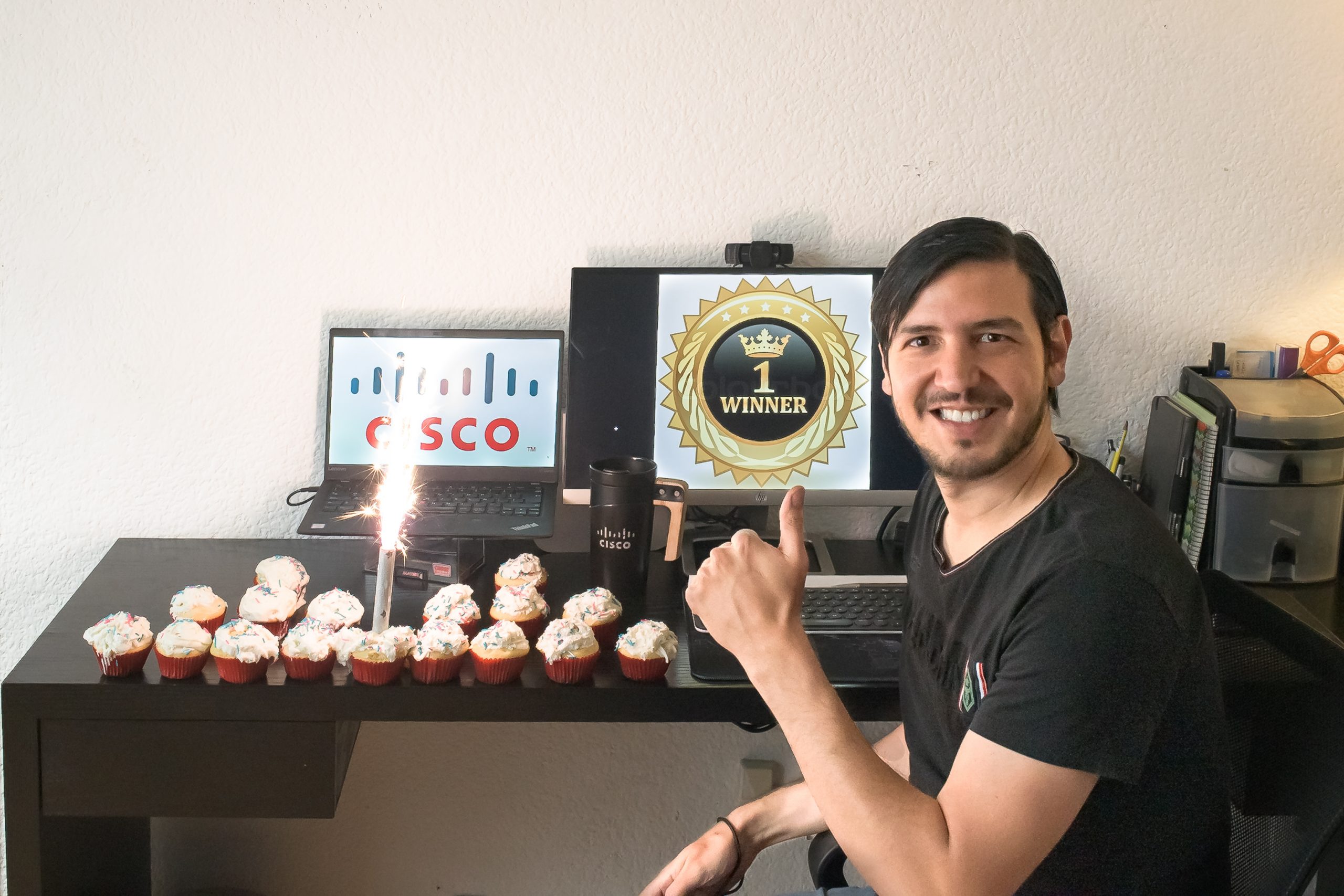 Javier sits at his desk with cupcakes in the shape of a Cisco logo and holds up his finger as #1.