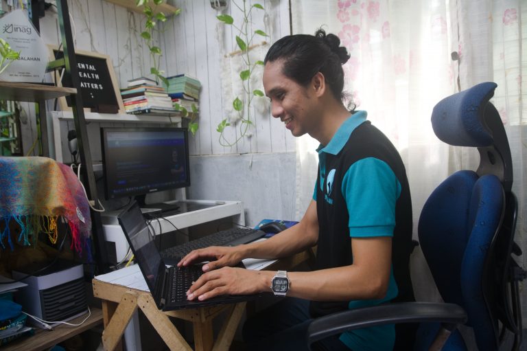 Ryan is wearing a teal and black polo shirt with Virtualahan logo. He is smiling while working on his laptop. He is in his workstation with an extra monitor, books, trophies, and hanging plants.