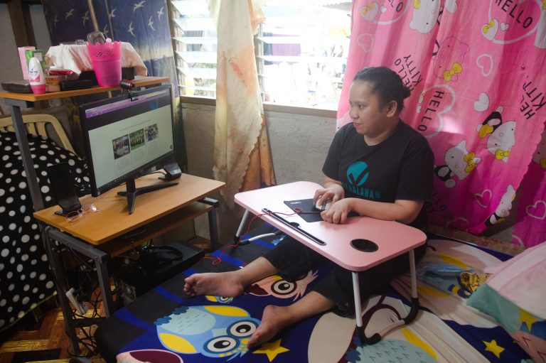 Grace is sitting in her bed while looking at her computer monitor and holding the mouse. She is wearing a black shirt with Virtualahan logo on it. She is in her room with a pink hello kitty curtain.