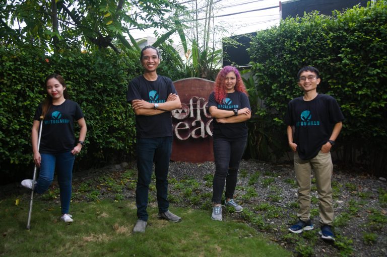 Four millennials (two females and two males) wearing black with Virtualahan logo on it striking a pose with green plants in the background.