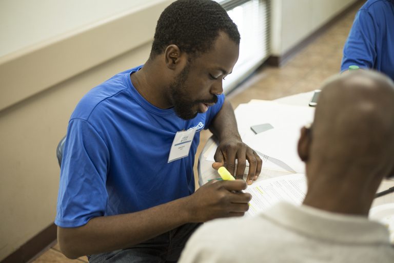 Man helping a homeless individual fill out a Mini City vital records application form. (Mini City + Community Bucket + Mercer Vital Records Outreach Event, Photo Credit: Anita Jones.)