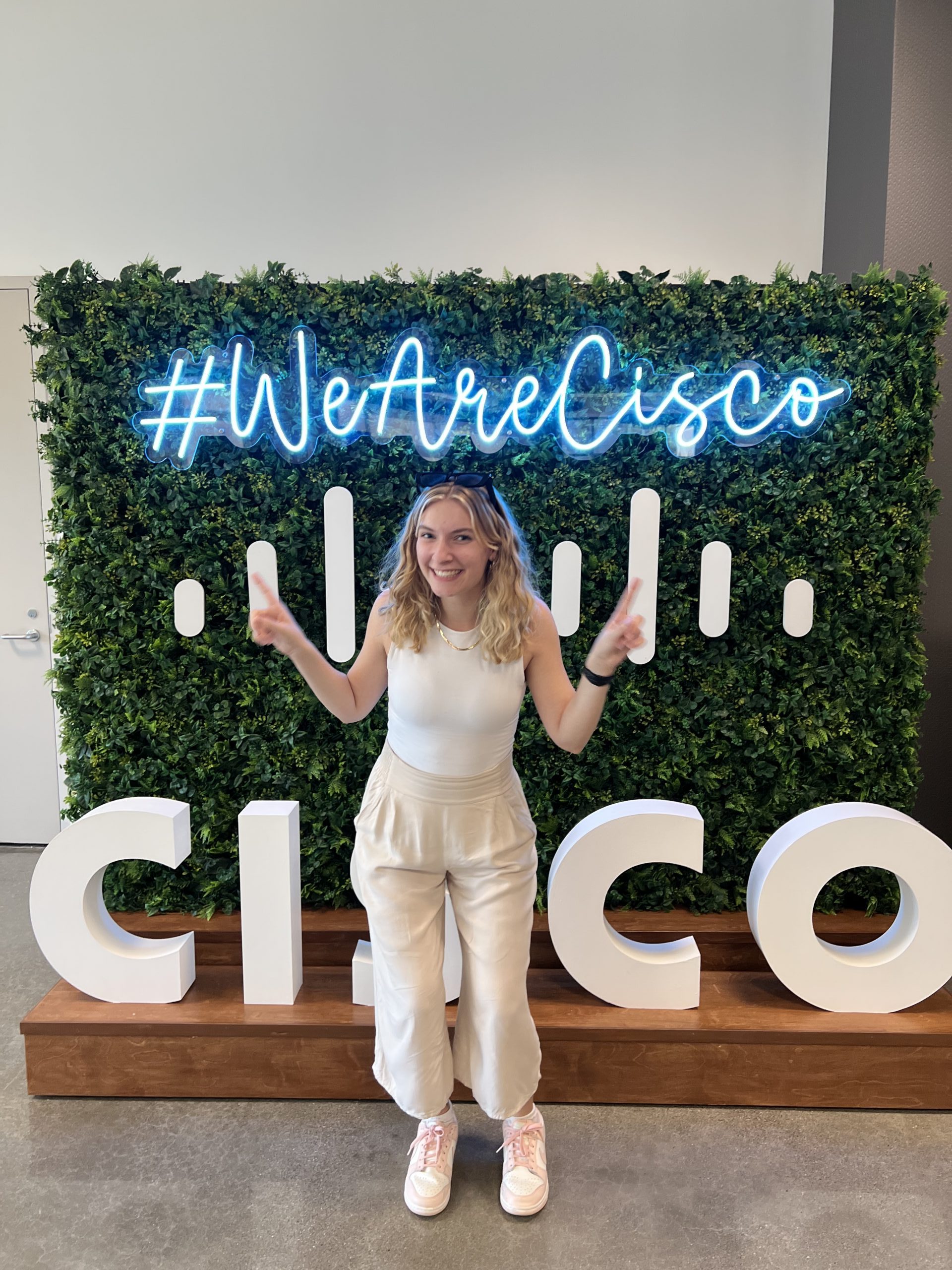 Young blond woman standing in front of neon #WeAreCisco sign with index fingers pointing up.