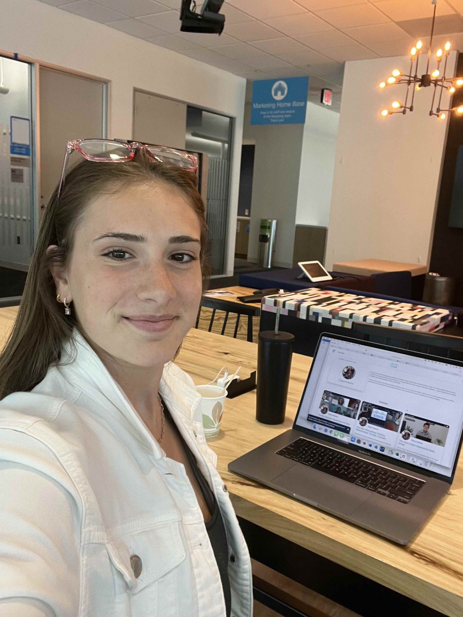 Young woman in white jacket takes selfie in front of laptop in office setting.