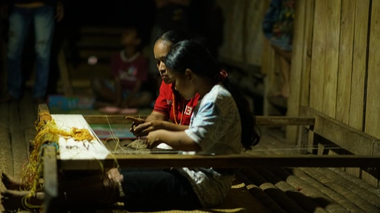 Two women weaving at night, with a light on above. 