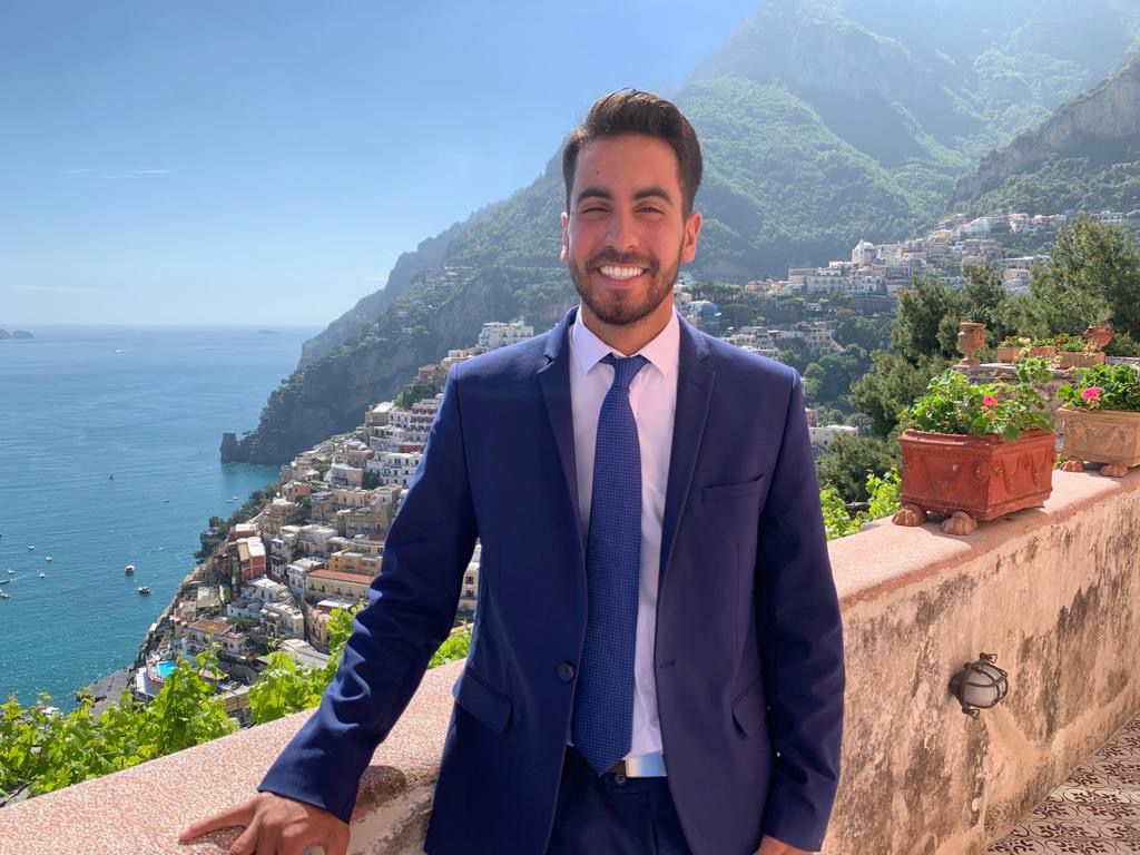 Tal standing against a wall with mountainous city seascape view behind him. 