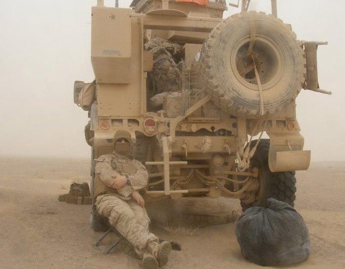 Travis, in military uniform, sitting behind a military vehicle.