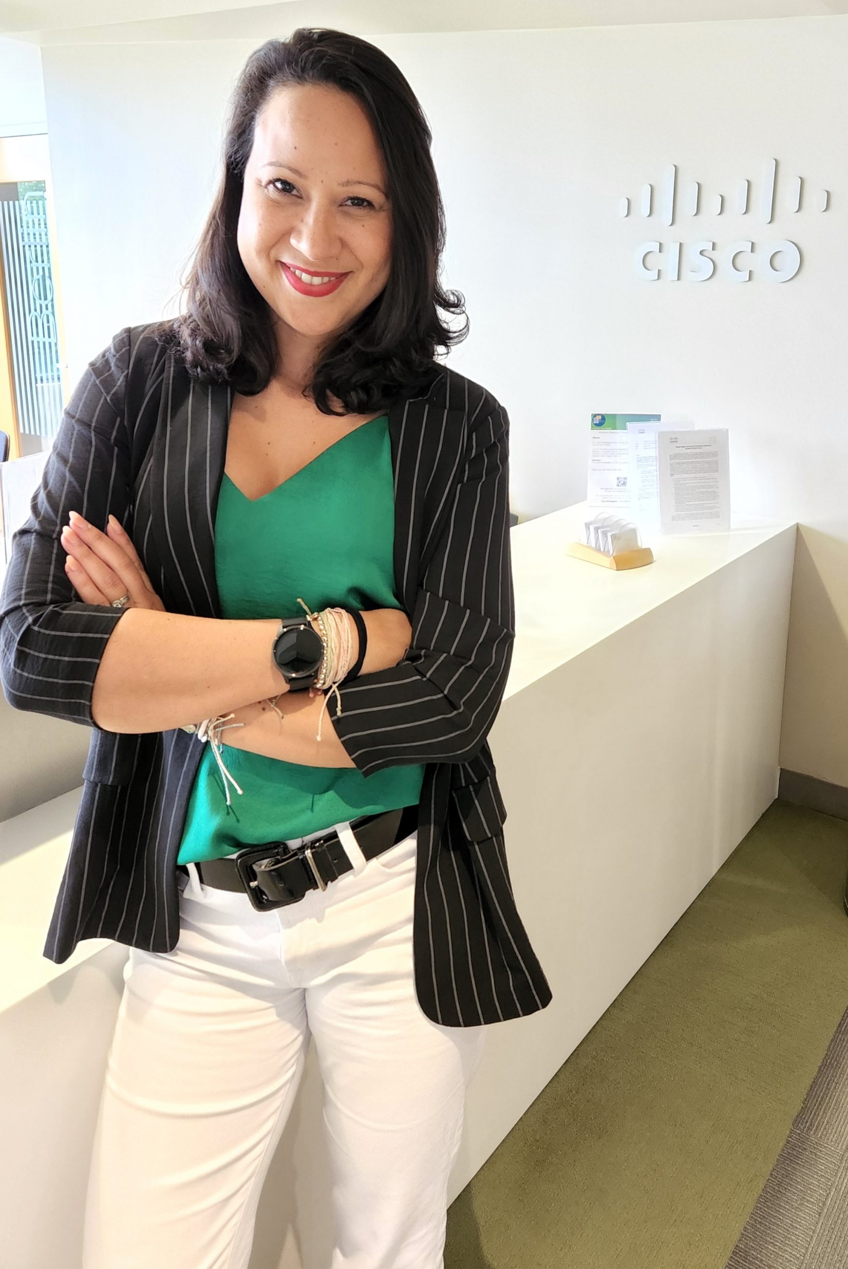 Suzi standing and smiling in foreground with arms crossed wearing black blazer and green blouse in Cisco office with Cisco logo behind her.
