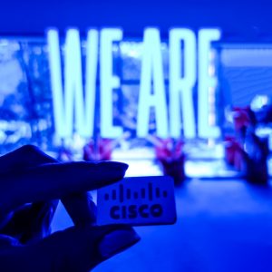 Hand holding Cisco logo tile in front of blue neon sign that reads "WE ARE"