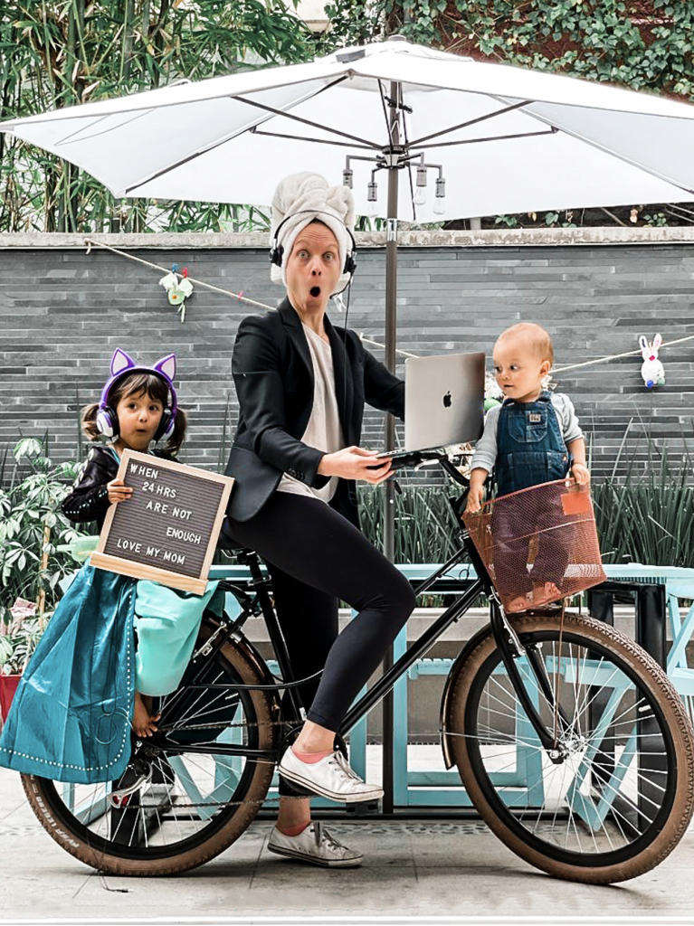 Woman wearing business suit and towel on her head, holding a laptop, and riding a bike with a child in the basket and another on the back. The child on the back is holding a sign that says, "When 24 hrs are not enough love my mom"