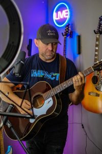 Man playing guitar wearing Cisco hat and Impact shirt.