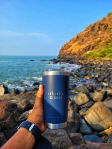 Blue and silver Cisco insulated coffee mug being held out with rocky seaside scene in the background.