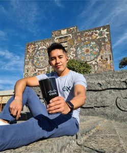 Man sitting on tiled rocks in Mexico holding insulated Cisco coffee mug out to the camera.