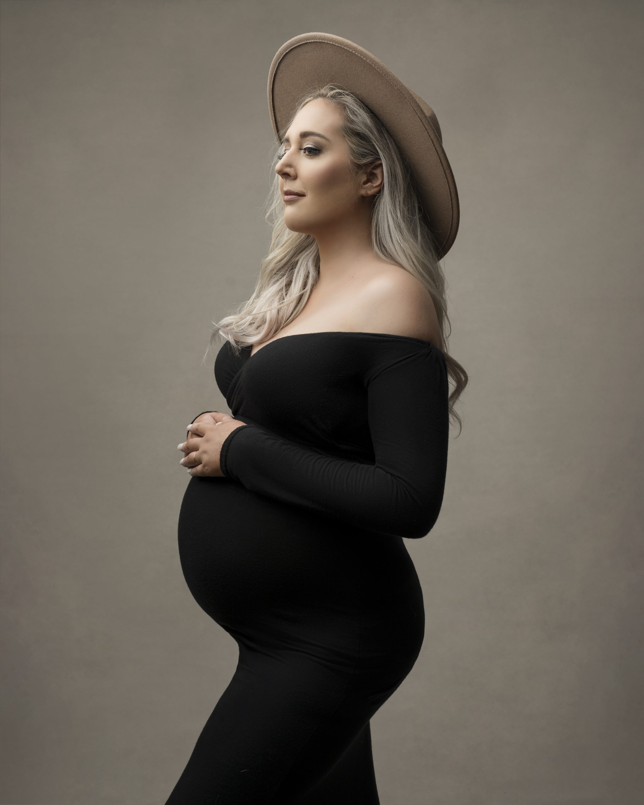 Allison wearing a black dress and beige hat while holding her baby bump.