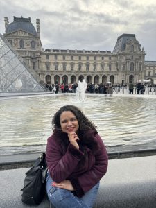 Mary, a brown woman with curly dark hair wears a burgundy coat with a fur collar. She smiles with her chin resting on her hand, the Louvre Museum and glass pyramid behind her.