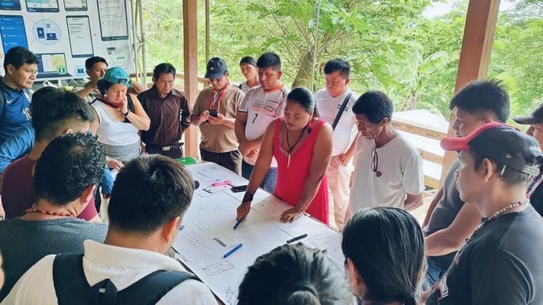 A large group of people looking at a map on a table together, with green trees behind them 