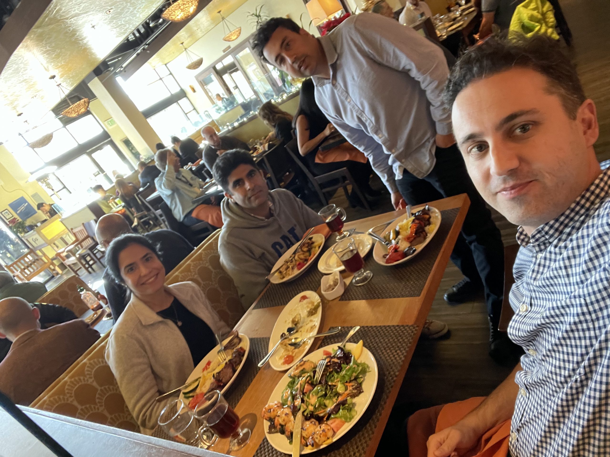 Niloo and her team sit at a table in a restaurant with plates of food on a table in front of them while one man takes a selfie