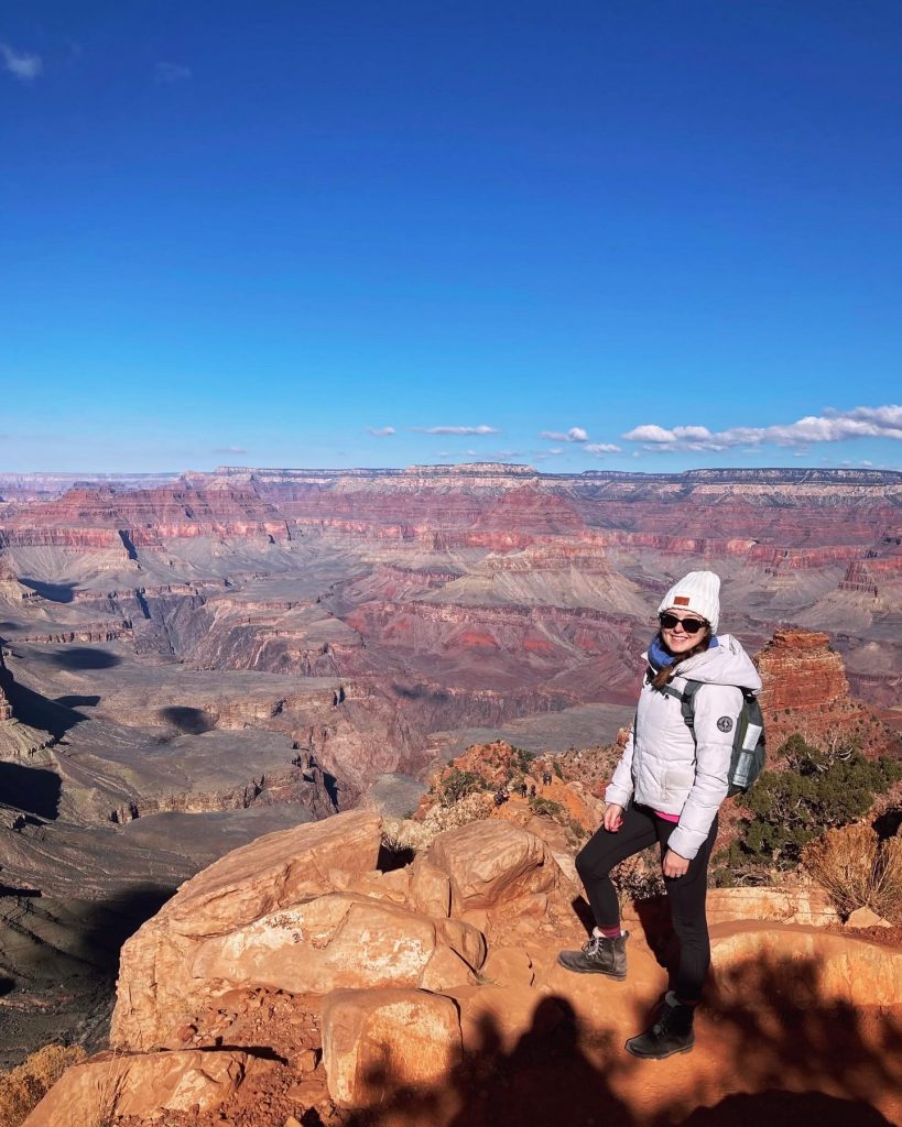 A woman wearing a white hat and jacket standing in front of the Grand Canyon.