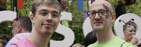 Two caucasian males wearing glasses, one in a pink t-shirt, one in a green t-shirt, standing in front of a big rainbow-logo Cisco pride parade float.