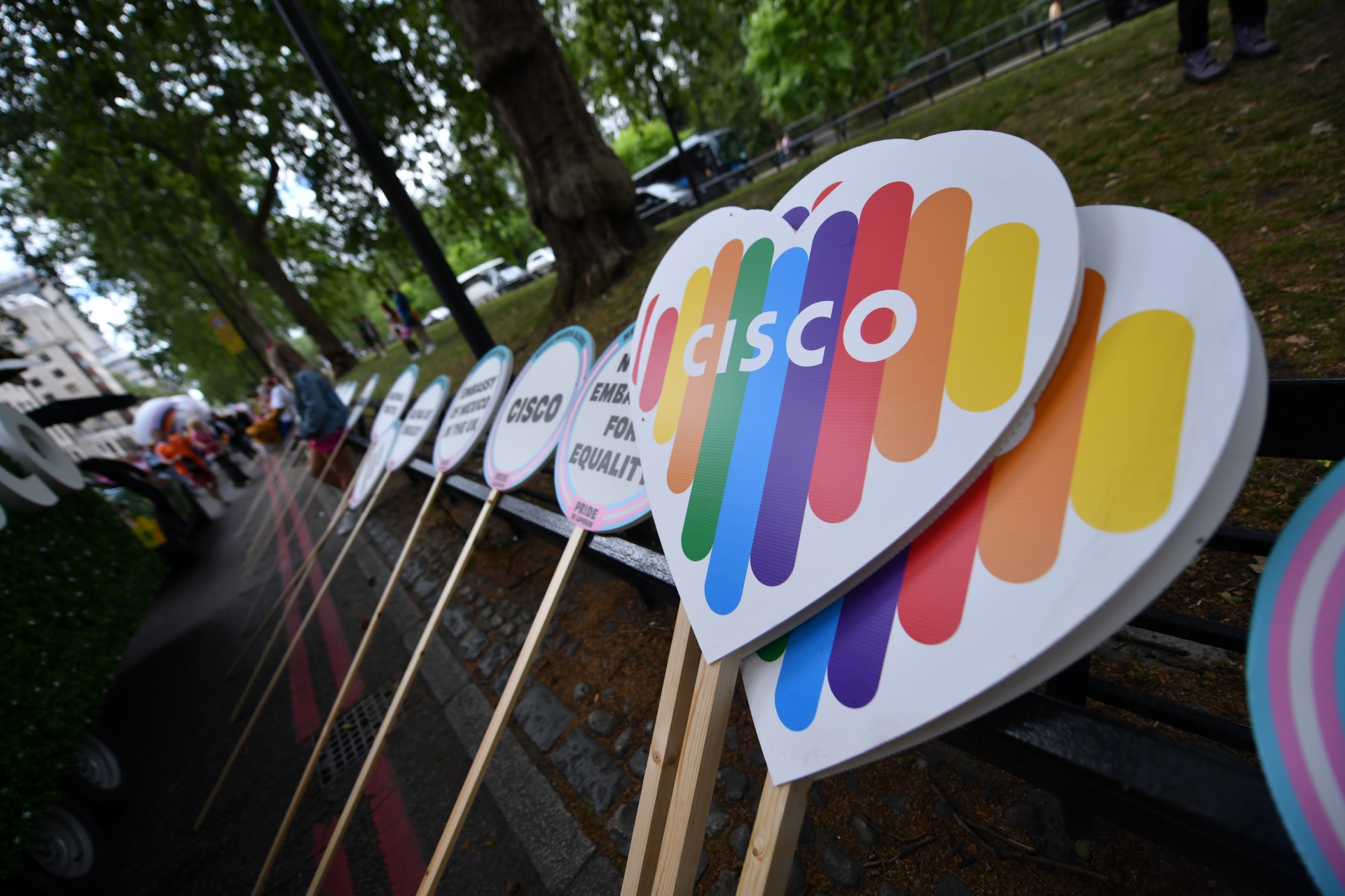 A stack of rainbow-colored Cisco pride logo signs, and other signs leaning on a fence in a park.