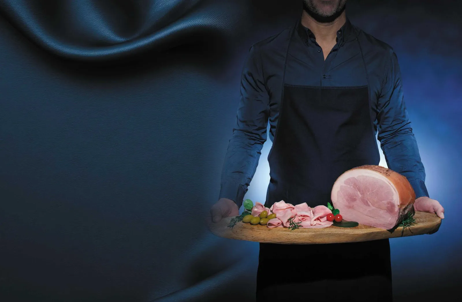 IMG-20260316-WA0002.webp Persona a mezzo busto che tiene in mano un tagliere con sopra il prosciutto cotto su sfondo blu