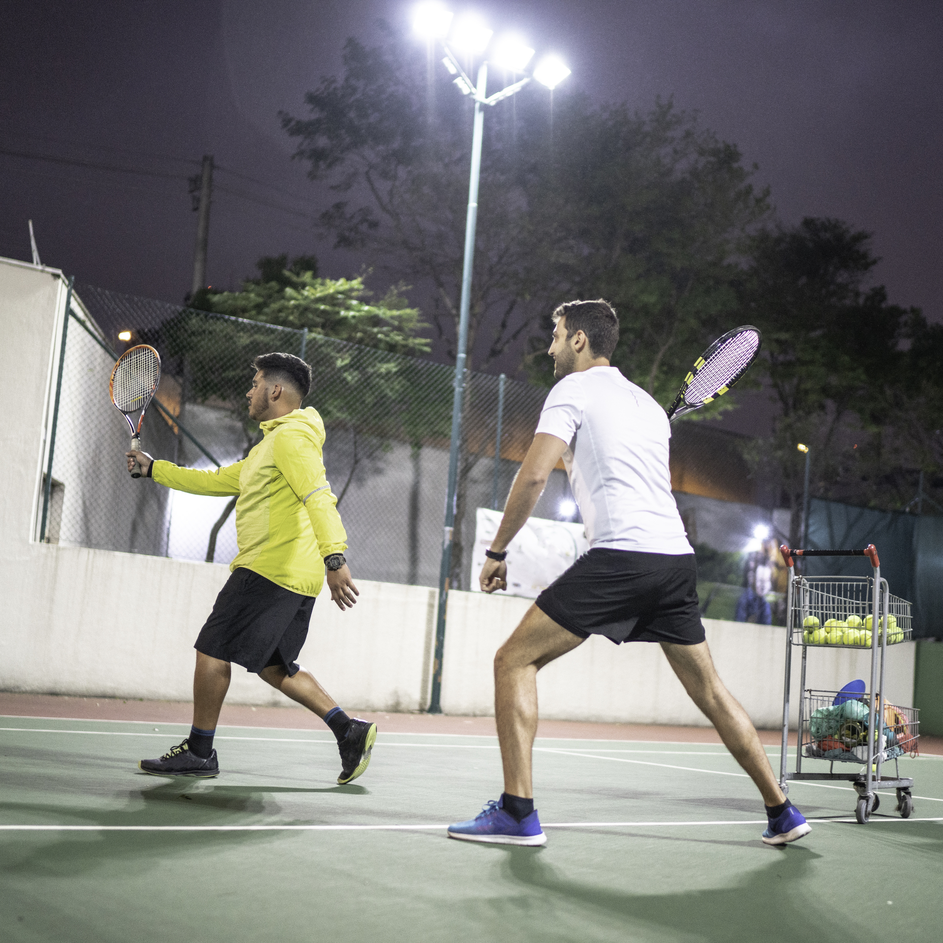 Doi bărbați joacă tenis pe un teren unde luminile s-au aprins automat pentru ora de joc programată.