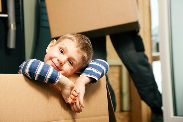 A smiling young child resting their head on their arms over the edge of a moving box.