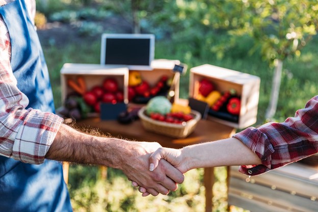 A closeup of a farmer shaking hands with another person with boxes of produce in the background.