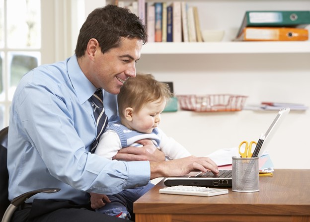 A person smiling and holding their child as they work at their desk from home.