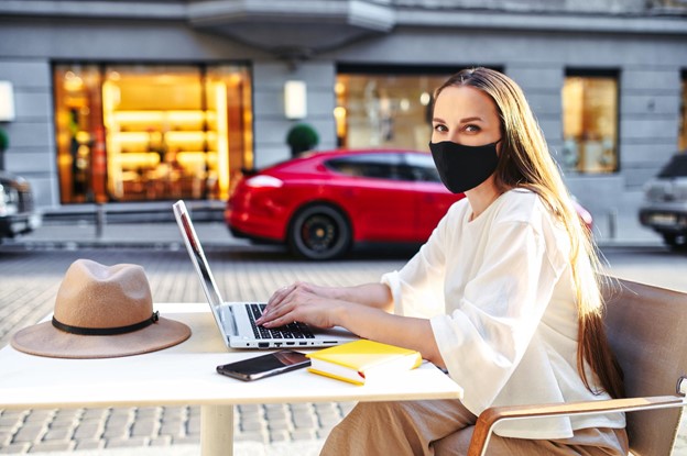 A person wears a facemask as they work on their laptop outdoors at a city cafe.