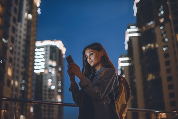 A young person using a smartphone on a city street at dusk, with buildings lighting up the skyline in the background.