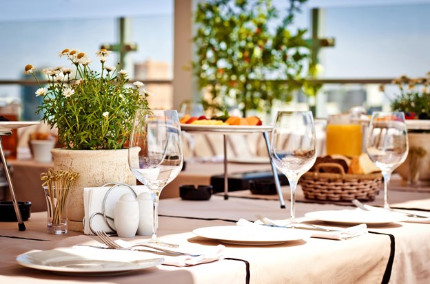 A table on the terrace of a restaurant is adorned with plates, wine glasses, and plants.