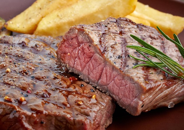 Close-up of a freshly grilled beef steak, topped with a sprig of rosemary and steak fries in the background.