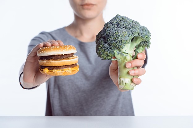 A closeup of a person holding an unhealthy hamburger in one hand and a healthier stalk of broccoli in the other.