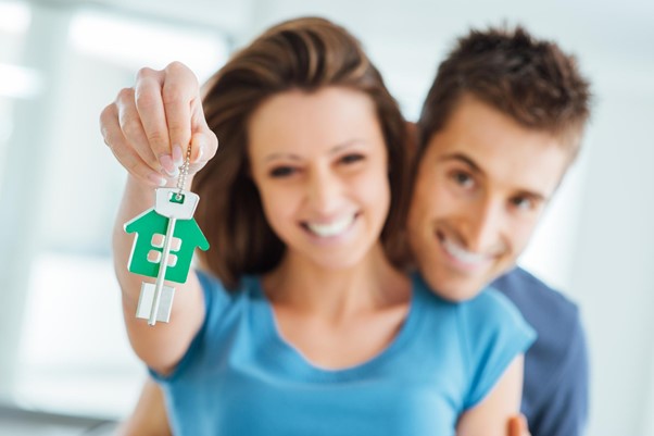 In the background, a smiling couple embraces, holding the key to their new house toward the foreground.