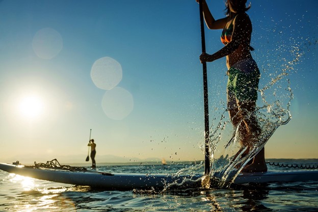 Two individuals standing atop their paddleboards on a lake, the sun shining in the background.