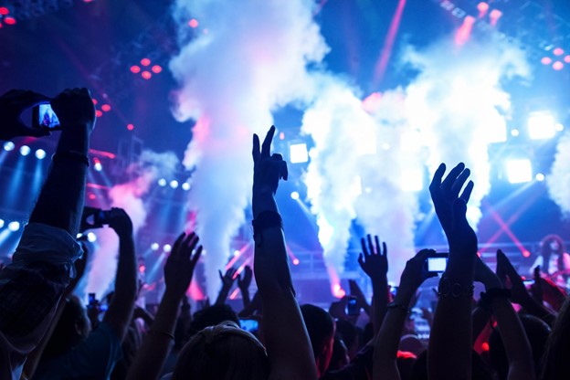 A crowd of raised arms with lights and a stage in the background of a dance club.