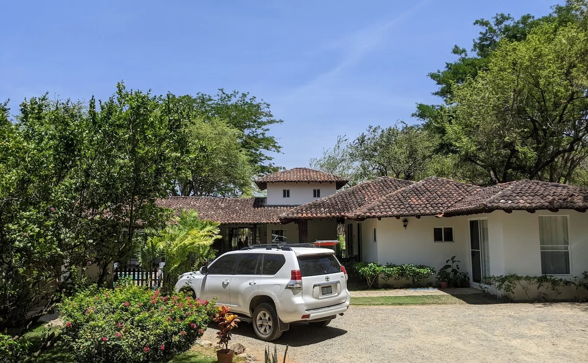 A tiled-roof house with a parked SUV, trees, flowering shrubs, and a bright driveway.