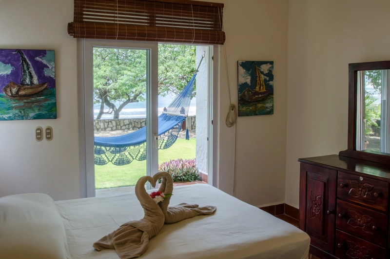 A cozy bedroom featuring a neatly made bed with towel art in the shape of a heart. The room is decorated with paintings of sailboats, and there's a wooden dresser with a mirror. A 