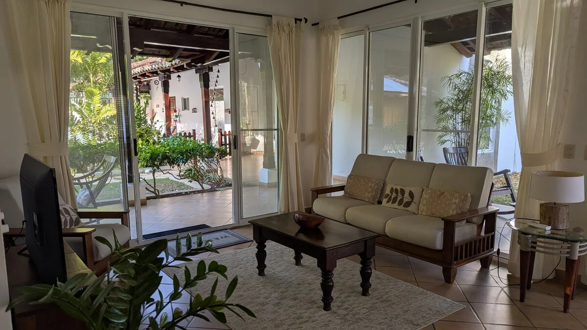A furnished living room with a sofa, coffee table, television, plants, and large glass doors opening toward a leafy outdoor area.