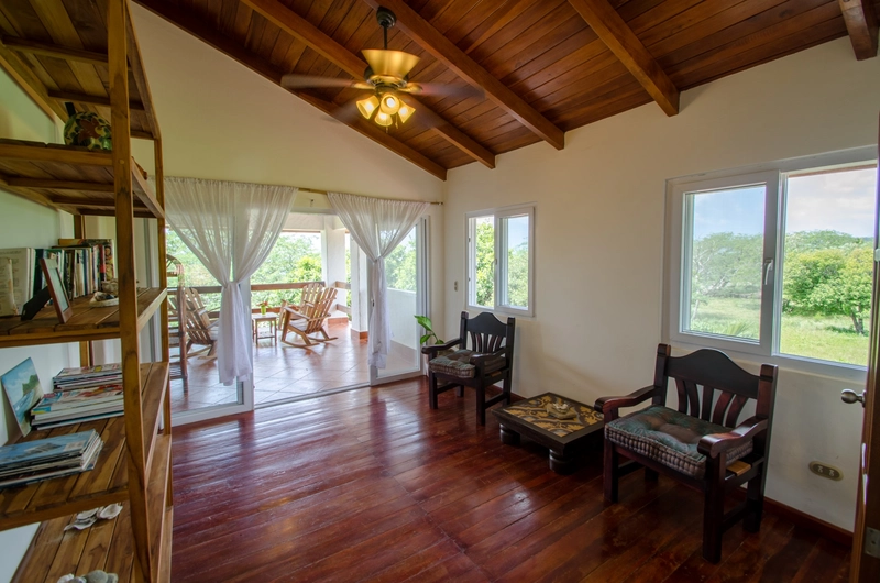 This image shows an interior room with a wooden ceiling and floor. There are two windows on the right side, and a set of chairs and a small table on the left. A bookshelf is visibl