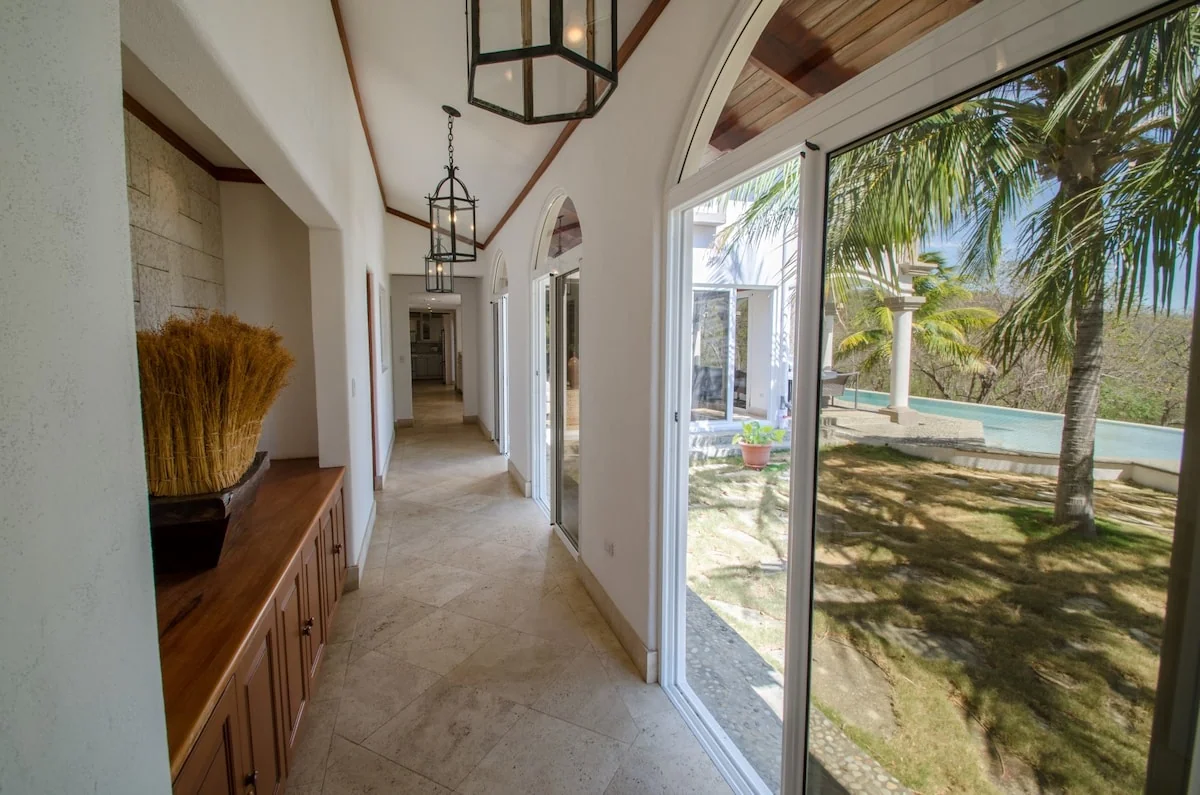 A bright arched hallway with hanging lanterns, a wooden cabinet, and large windows overlooking palm trees and a pool.