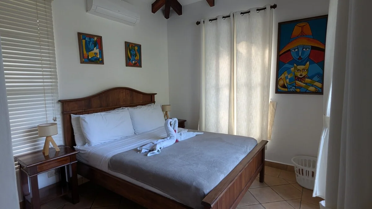 A tidy bedroom with a wooden bed, gray bedding, white curtains, framed colorful artwork, and bedside lamps.