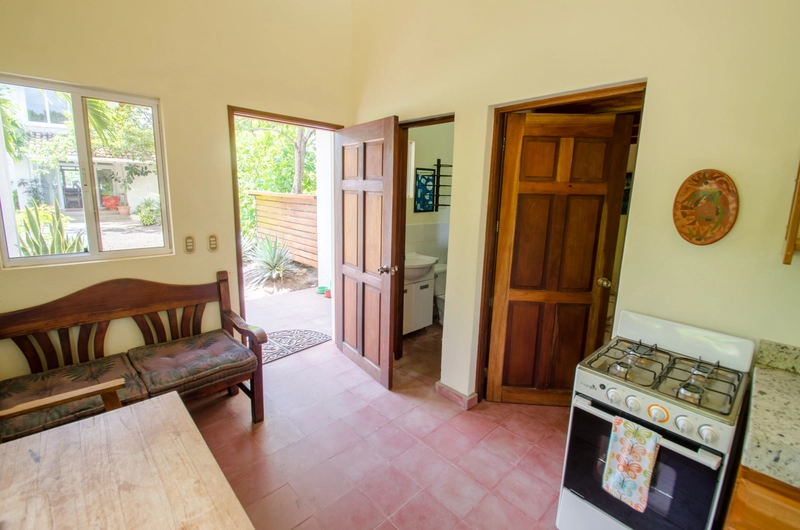 The image shows a cozy kitchen with natural light illuminating the space. It features wooden doors, one of which is open and offers a view of a lush outdoor area with plants. Insid