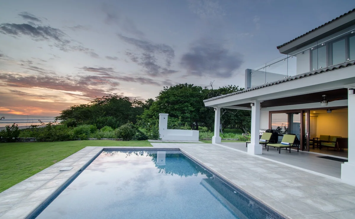 A rectangular swimming pool beside a modern white house opens onto a tiled patio, lawn, trees, and a sunset sky.