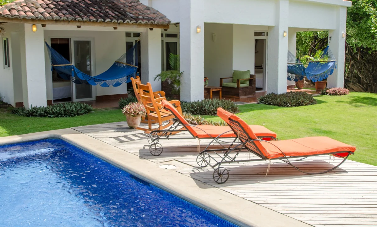 A bright poolside area features orange loungers, blue hammocks, a white house, and manicured lawn.