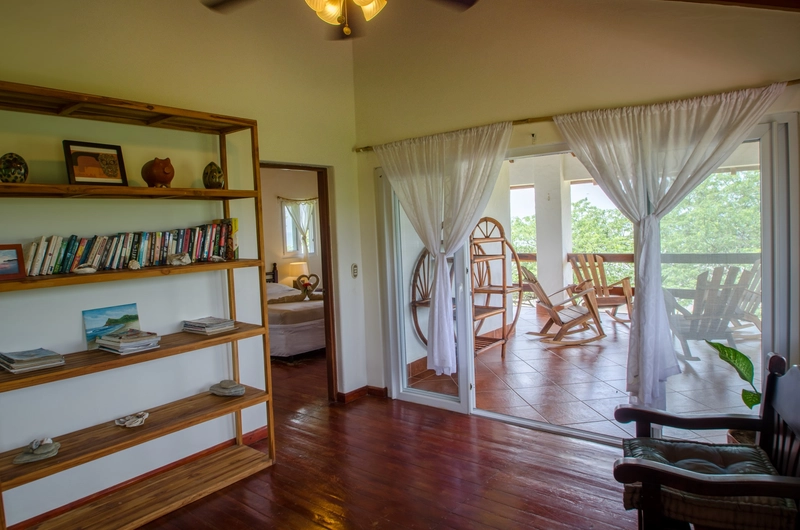The image depicts a cozy interior space featuring a prominent wooden bookshelf filled with books and decorative items on the left. To the right, white sheer curtains partially cove